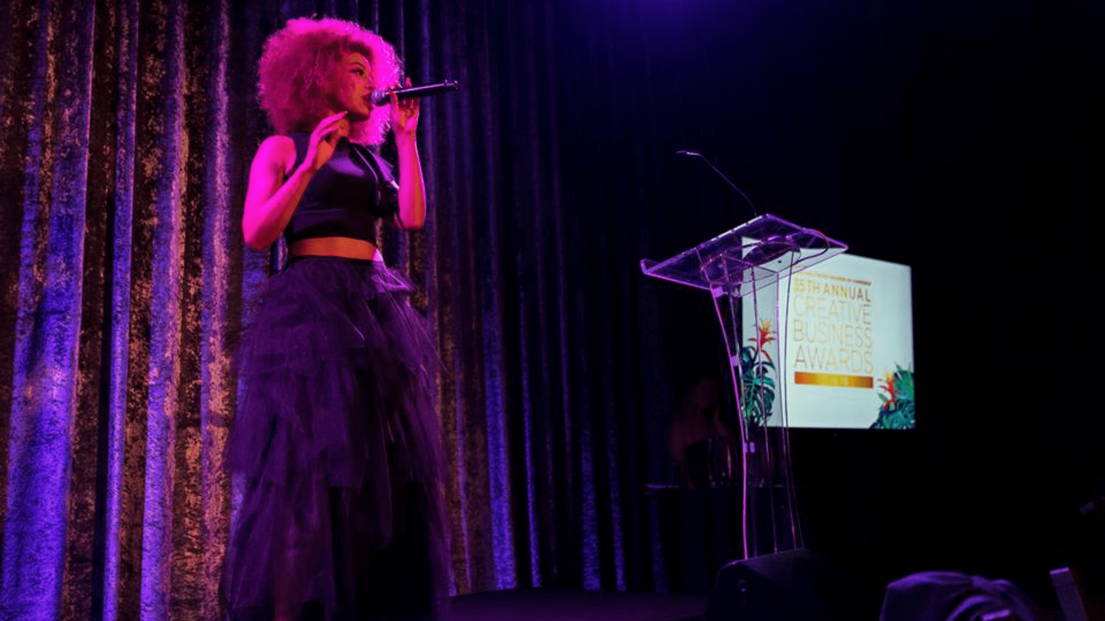 A female singer performing live at an award show, illuminated by pink and purple stage lighting, holding a microphone.