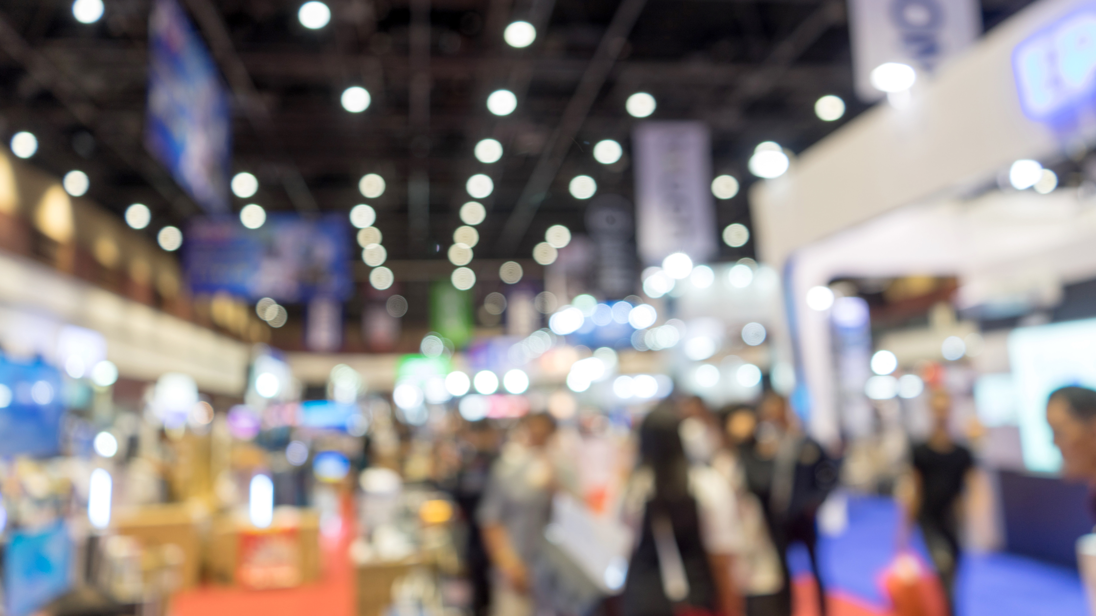 A blurred image of a bustling trade show with bright lights, attendees walking around, and booths to explore.